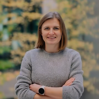 Eine lächelnde Frau in grauem Strickpullover mit verschränkten Armen vor Herbstlaub