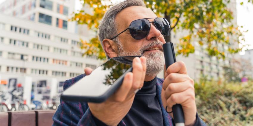 A blind man sits on a bench outdoors and listens to the voice output of his smartphone.