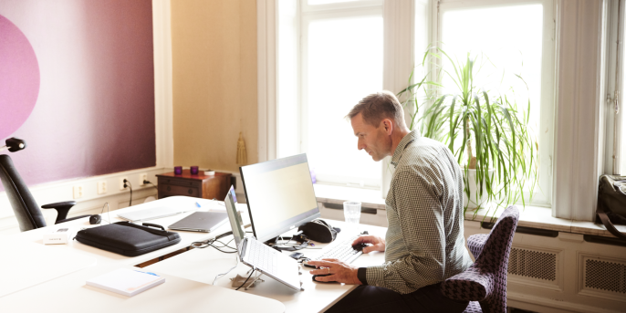 A man sits at a writing table. In front of him are a screen, laptop and other work equipment.