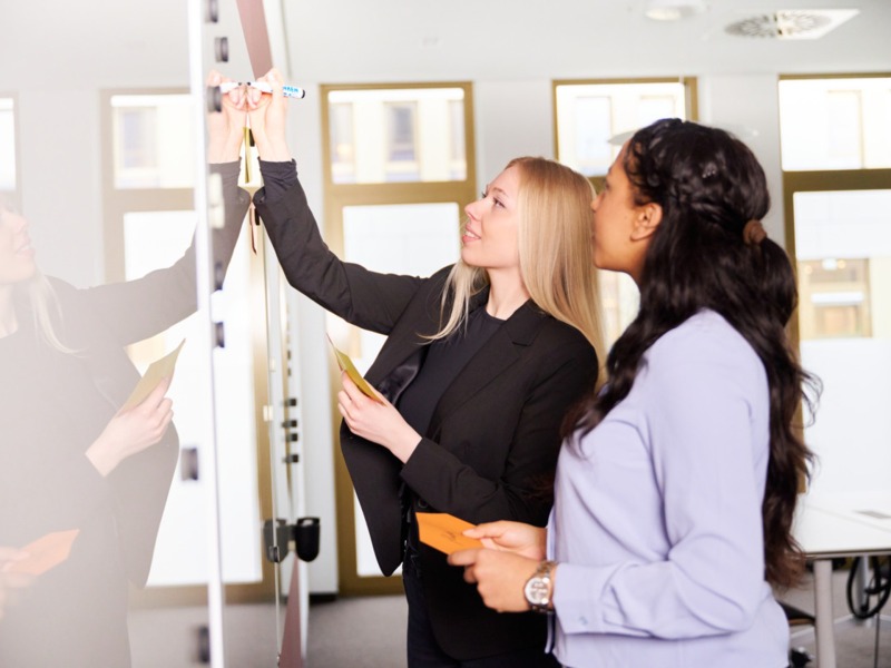 Zwei Frauen stehen an einem Whiteboard und bringen daran Notizen für den Workshop an.