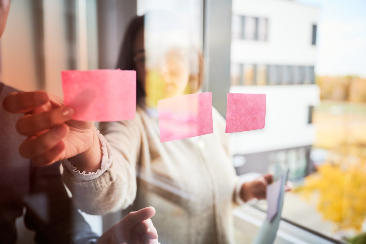 Mitarbeitene kleben Post its an eine Glaswand Mitarbeitene kleben Post its an eine Glaswand