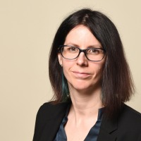 Business-Portrait von Nina Gerling mit halblangem braunem Haar und grünen Strähnen, freundlich in die Kamera lächelnd. Sie trägt eine schwarze Brille, einen schwarzen Blazer und eine glänzende schwarze Bluse.